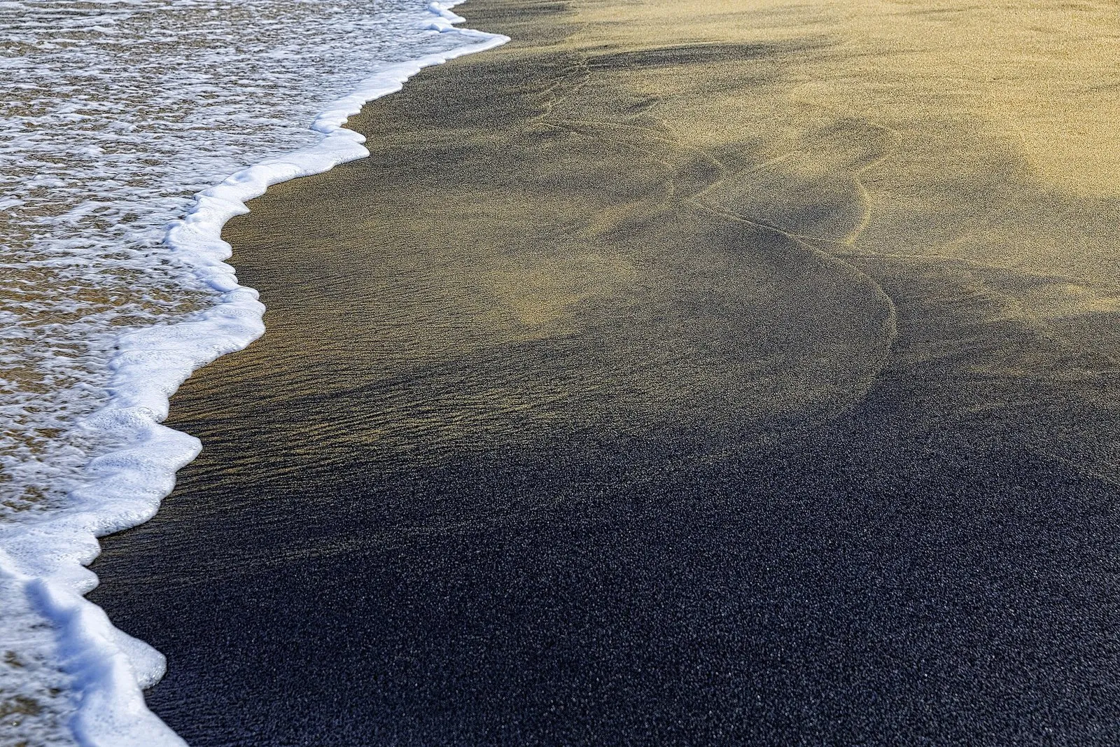 Faixa de areia escura, quase preta, na beira do mar, com a espuma de uma onda à esquerda e areia mais clara ao fundo