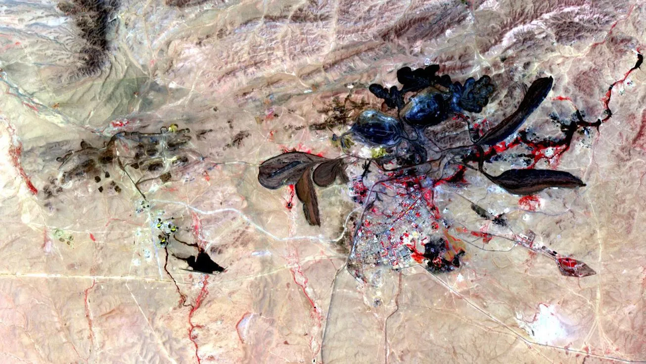Imagem de satélite em cores falsas de uma região árida, com grandes cavas de mineração escuras, lagos de rejeitos alongados e uma cidade ao centro
