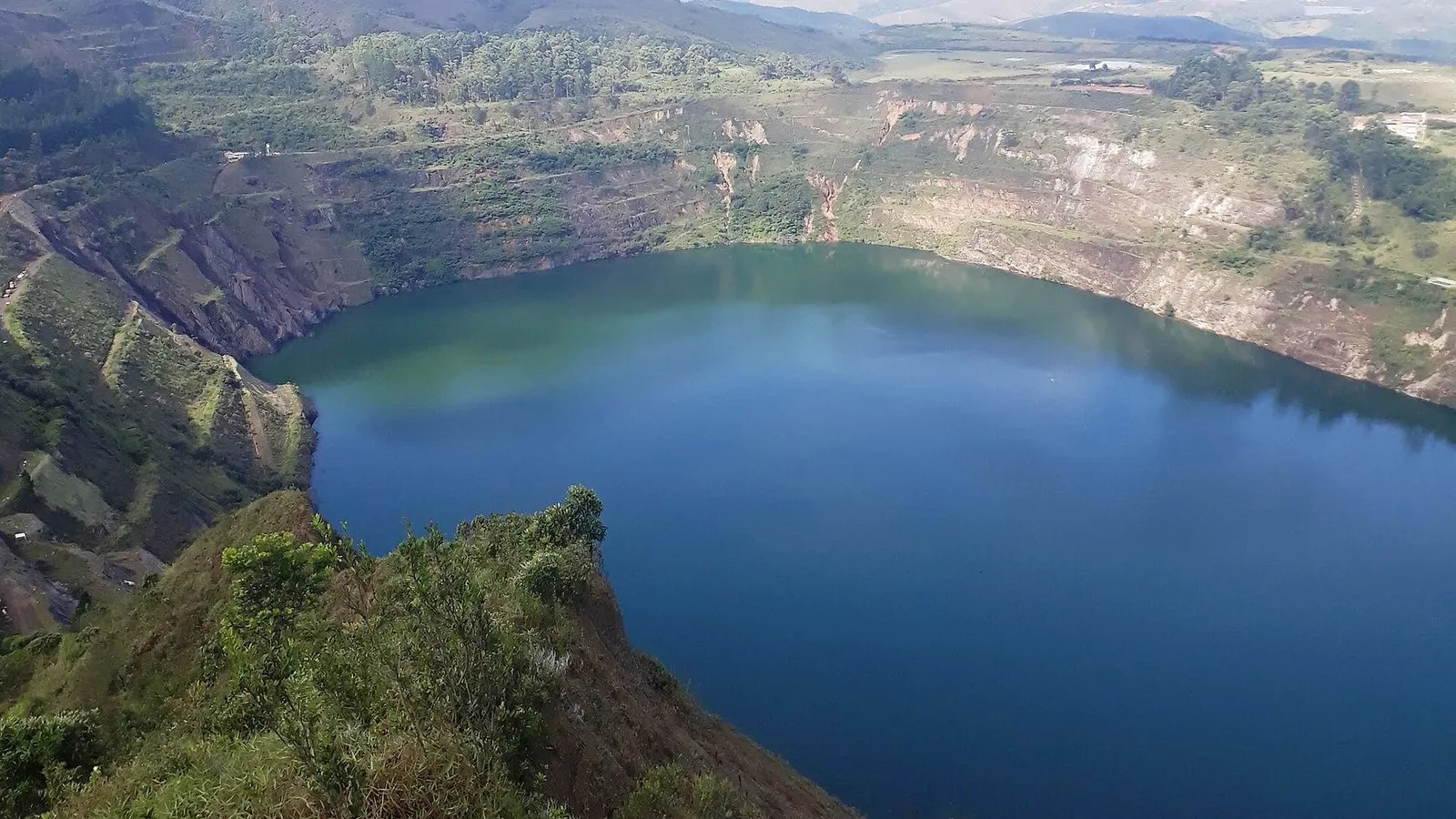 Cava de mineração a céu aberto ocupada por um grande lago azul, com paredes em degraus parcialmente cobertas de vegetação e morros ao fundo