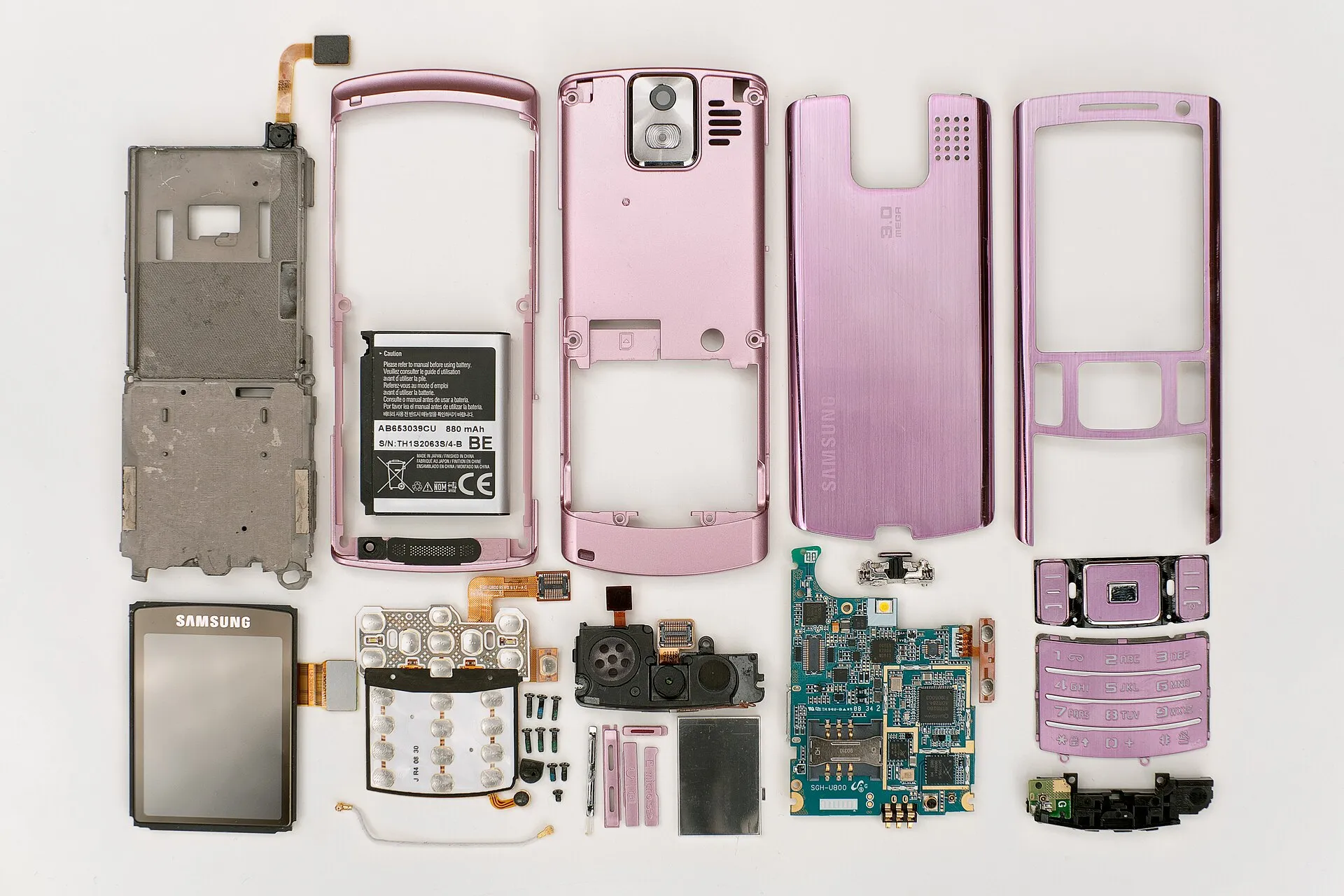 Peças de um celular antigo desmontado sobre fundo branco: carcaças metálicas rosadas, bateria, tela, teclado, placa de circuito e pequenos parafusos