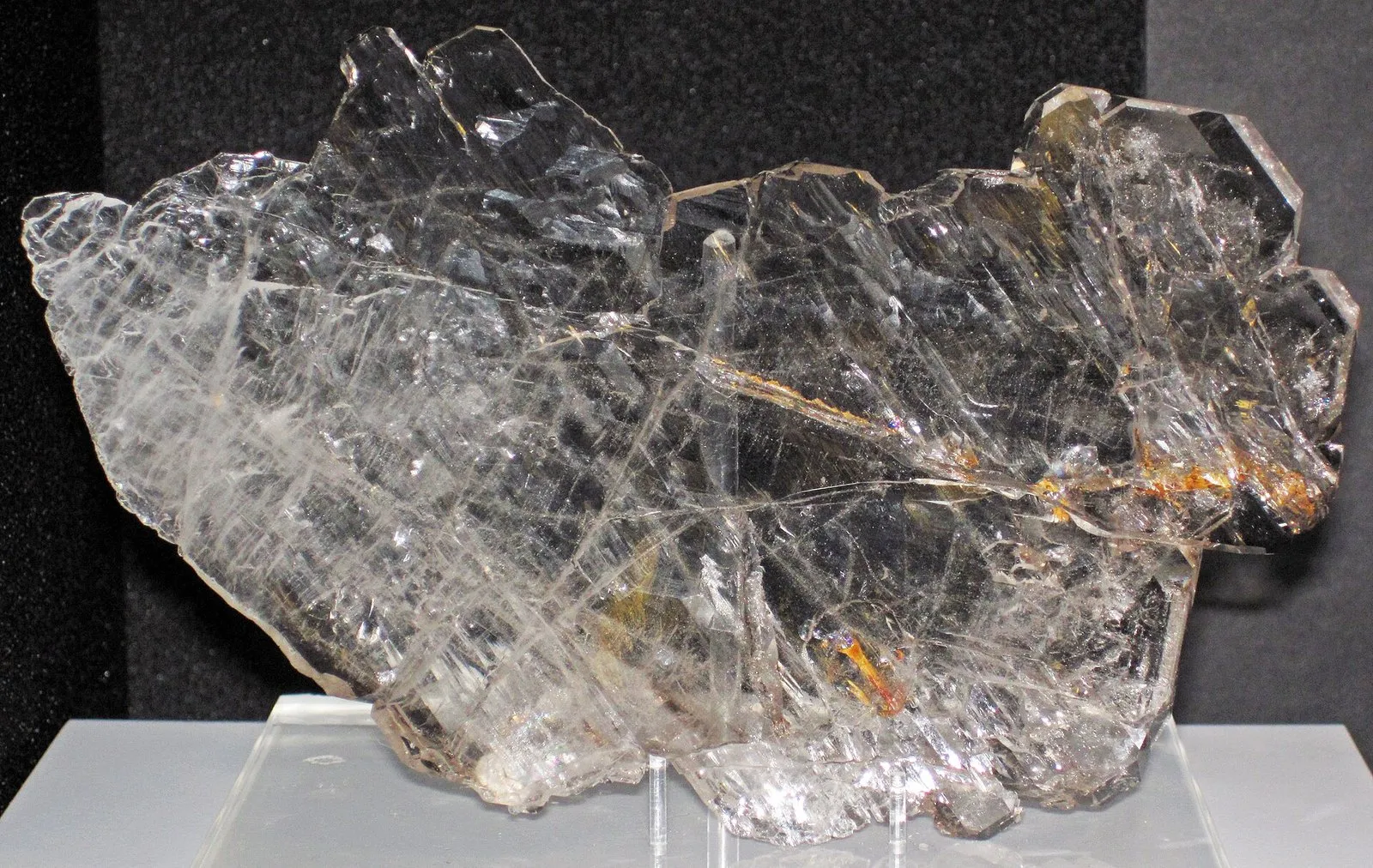 Cristal de quartzo transparente e incolor, com faces planas e algumas inclusões alaranjadas, sobre um suporte de acrílico e fundo preto