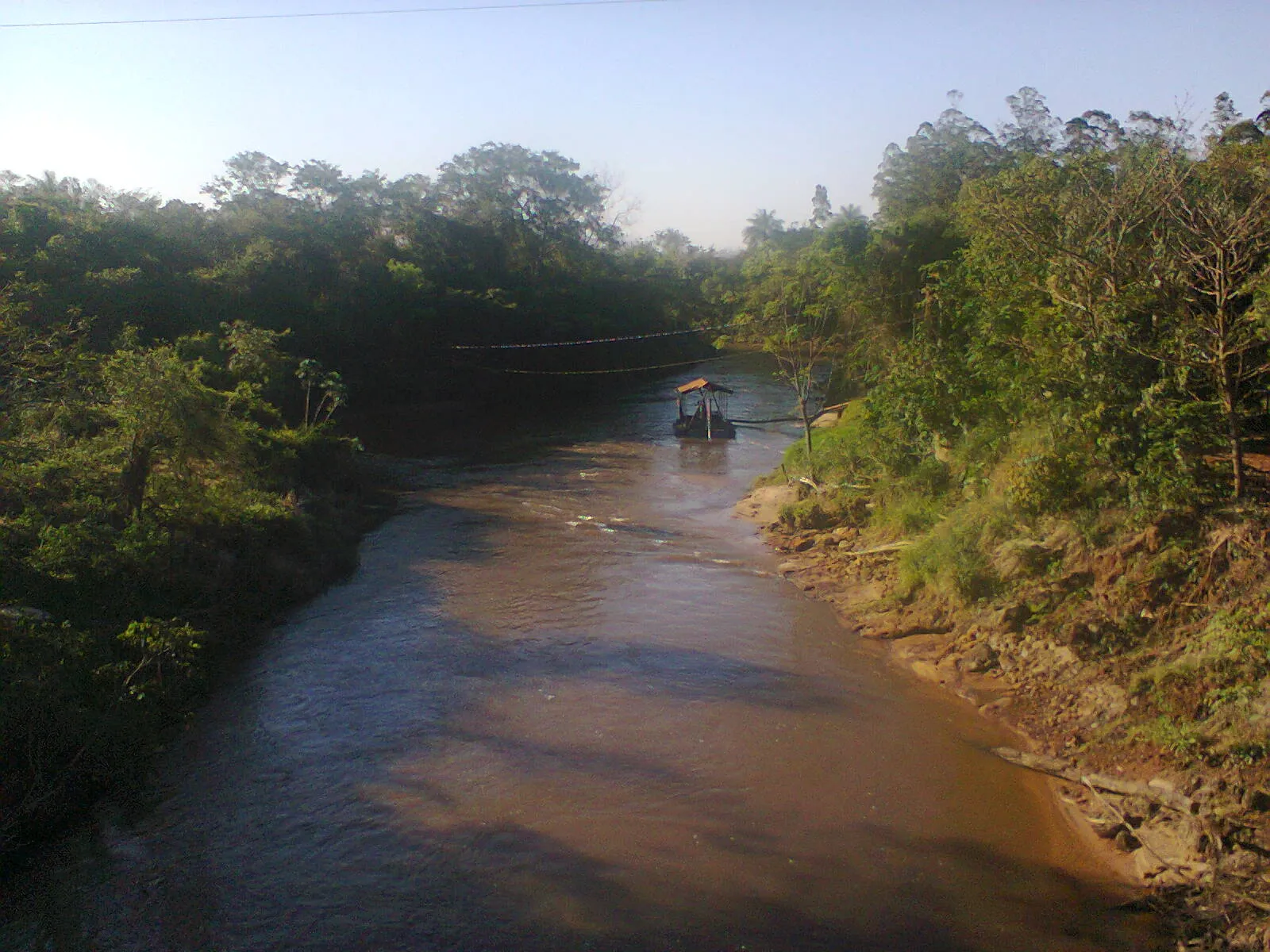 Rio de águas barrentas cercado de mata, com uma pequena draga de areia flutuando ao fundo e um cabo atravessado sobre a água