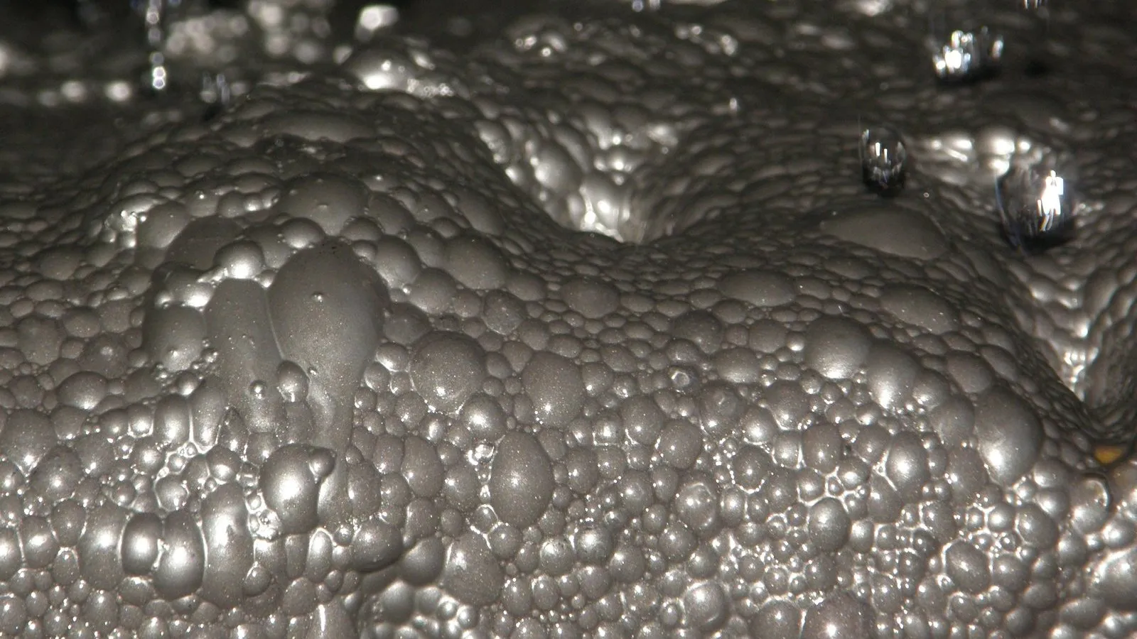 Close de uma espuma cinza-metálica formada por bolhas de tamanhos variados, carregadas de partículas minerais finas