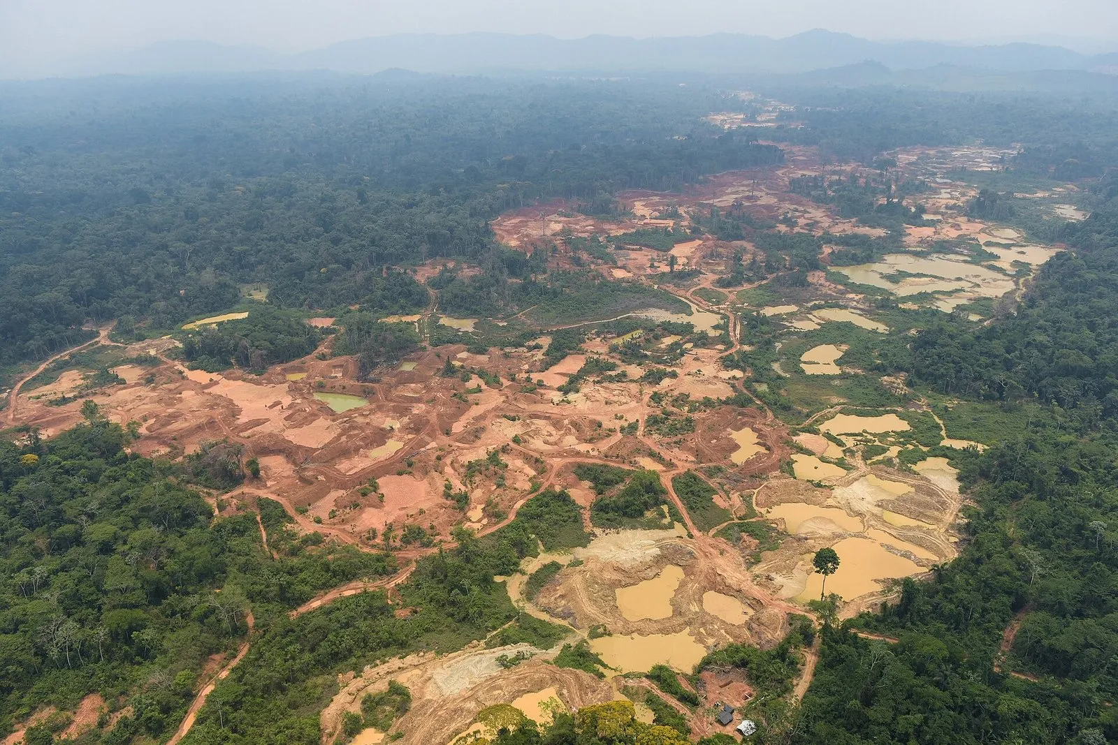 Vista aérea de uma grande área de floresta desmatada pelo garimpo, com solo avermelhado revirado, poças de água barrenta e estradas de terra, cercada por mata