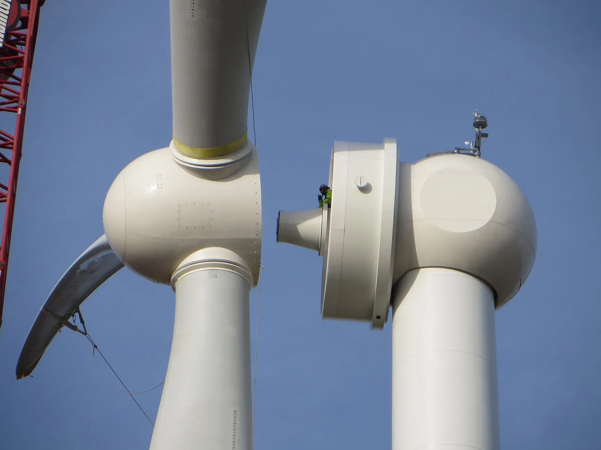 Turbina eólica branca em montagem, com o cubo e as pás suspensos ao lado da nacele, onde aparece o grande anel do gerador