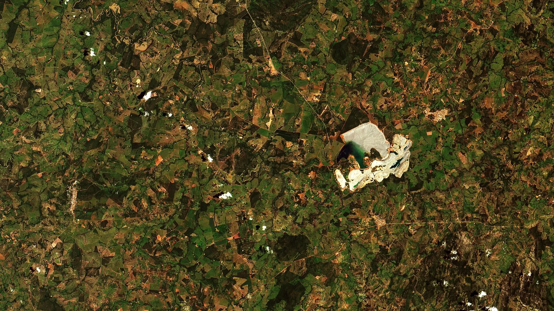 Imagem de satélite de uma mina a céu aberto em meio a áreas de pasto e vegetação, com a cava e o reservatório em tons de branco e verde