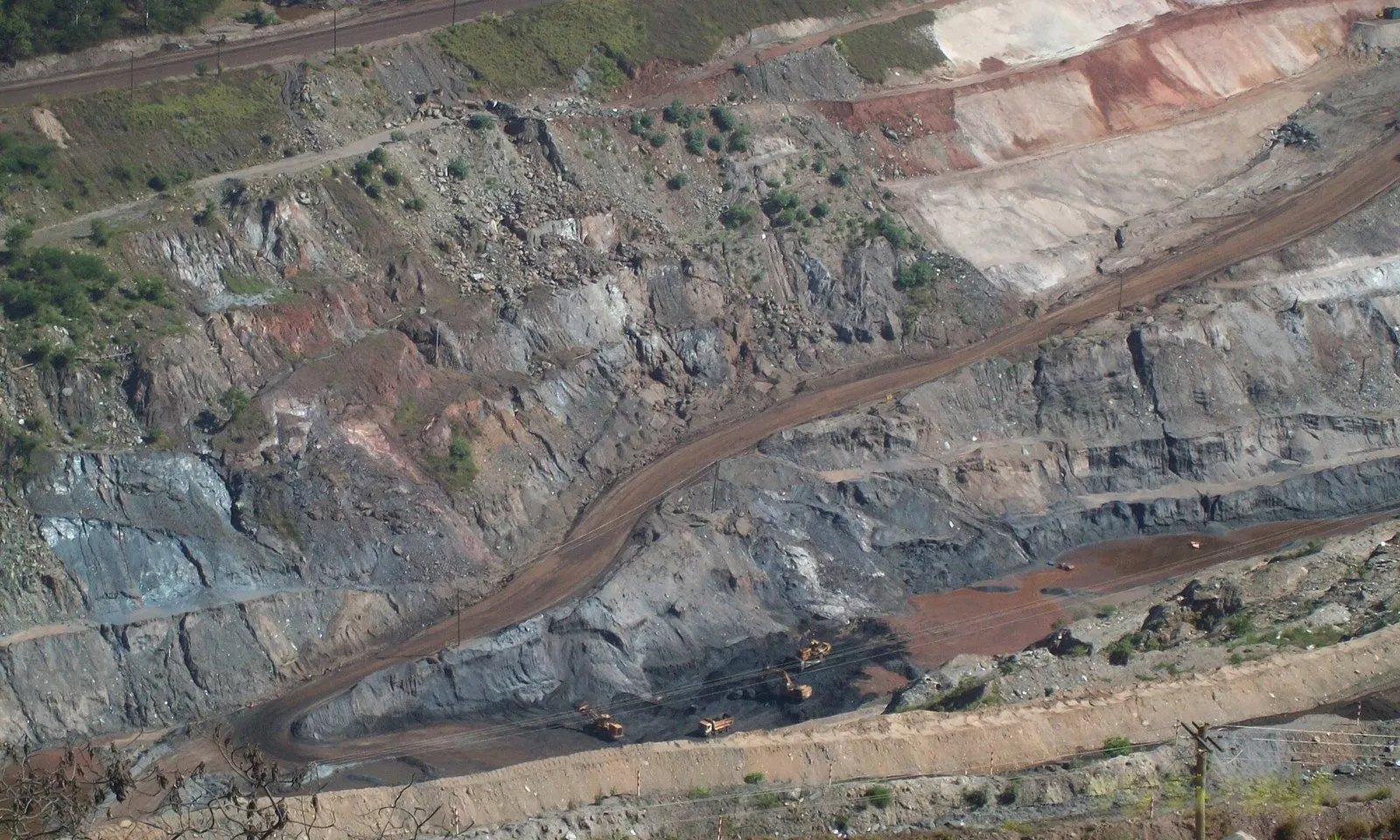 Cava de mina de ferro vista do alto, com paredes de rocha cinza e avermelhada, uma estrada de terra em curva e máquinas de carga no fundo