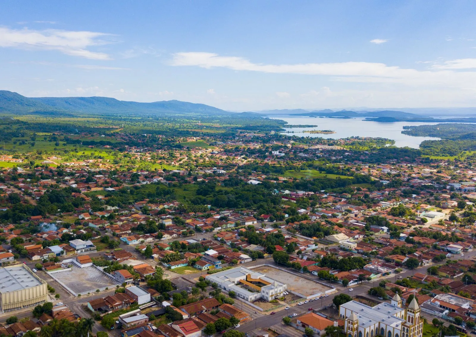 Vista aérea de uma cidade com casas de telhado vermelho e ruas arborizadas, com morros e um grande lago ao fundo