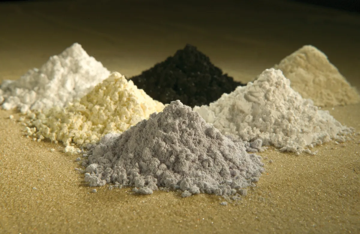 Seis montes de pó de óxidos de terras raras, em tons de branco, bege, cinza e preto, sobre uma superfície de areia