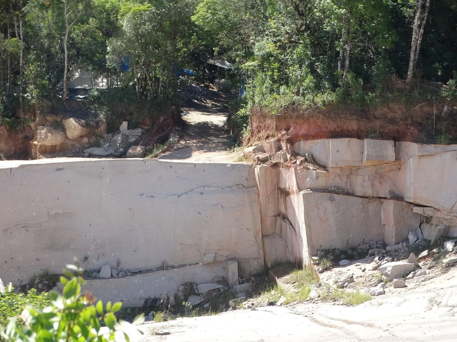 Paredão de granito claro cortado em blocos numa pedreira, com vegetação e terra avermelhada no alto