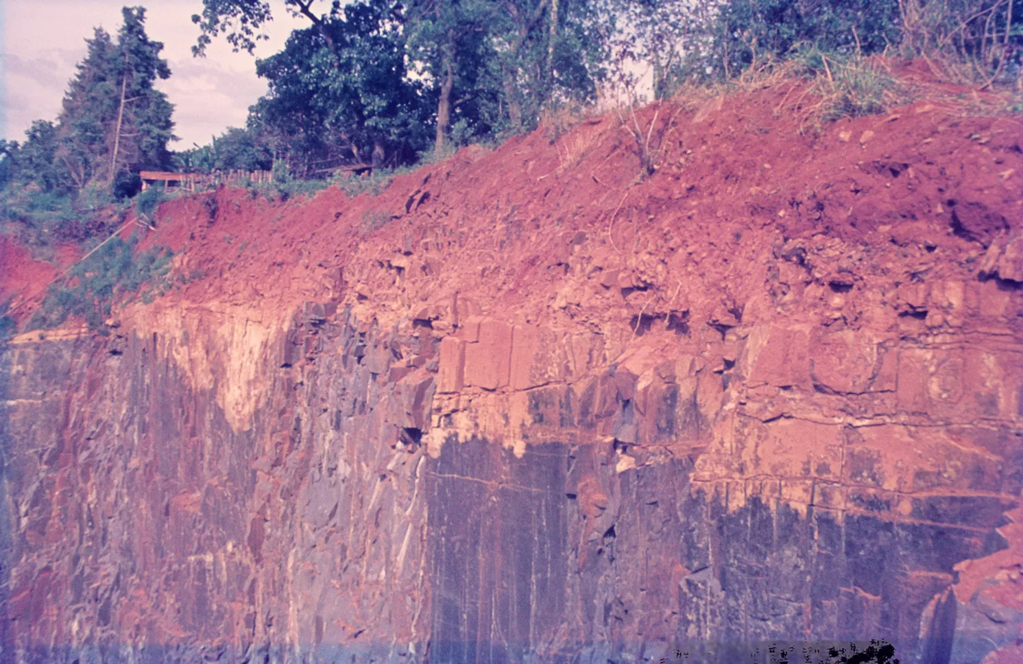 Corte de terreno com solo vermelho no topo, uma faixa intermediária mais clara e rocha escura fraturada embaixo, com árvores ao fundo
