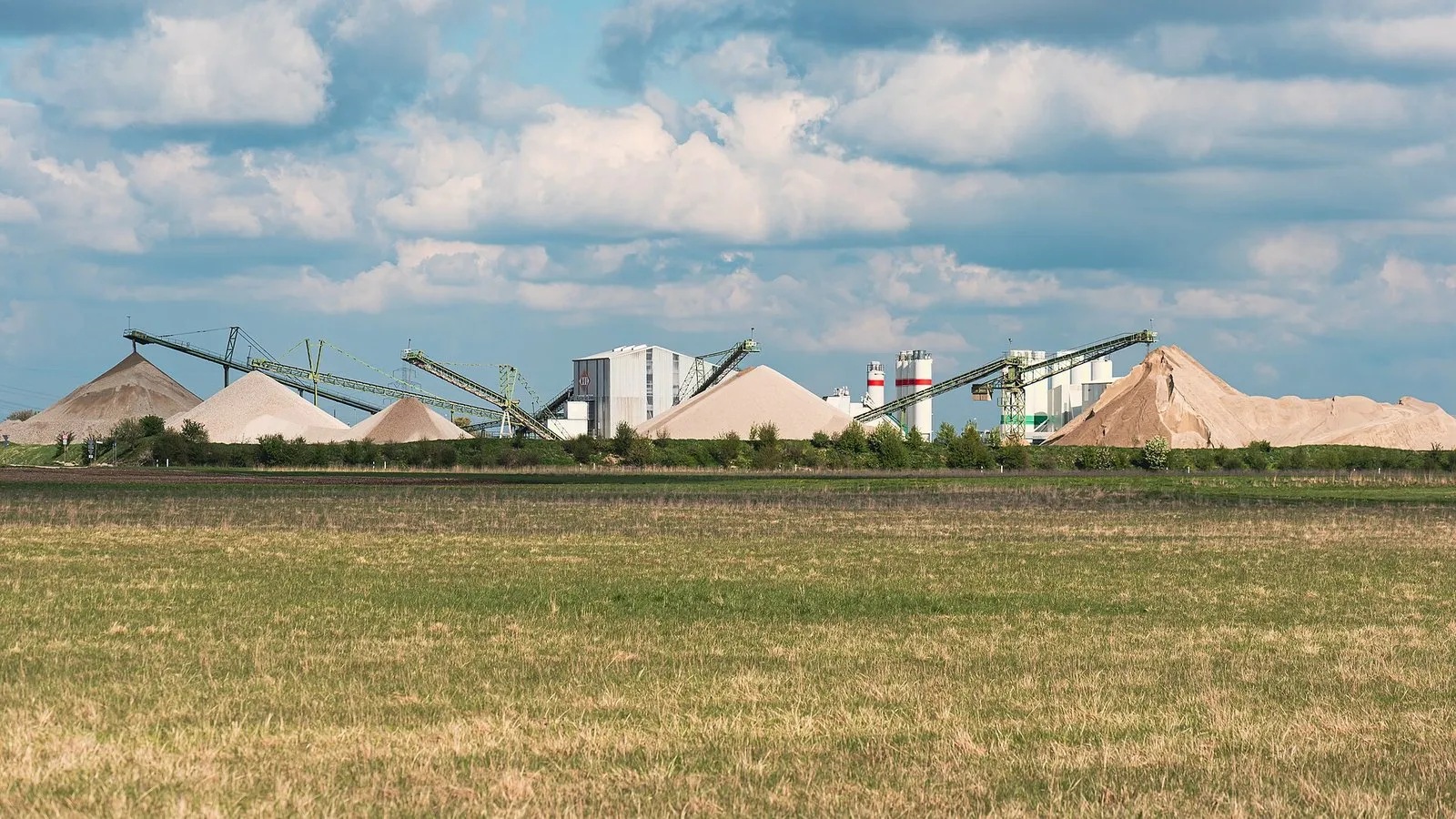 Grandes pilhas cônicas de areia e cascalho, esteiras transportadoras elevadas, silos e galpões de uma mineradora de agregados, vistas de longe através de um campo
