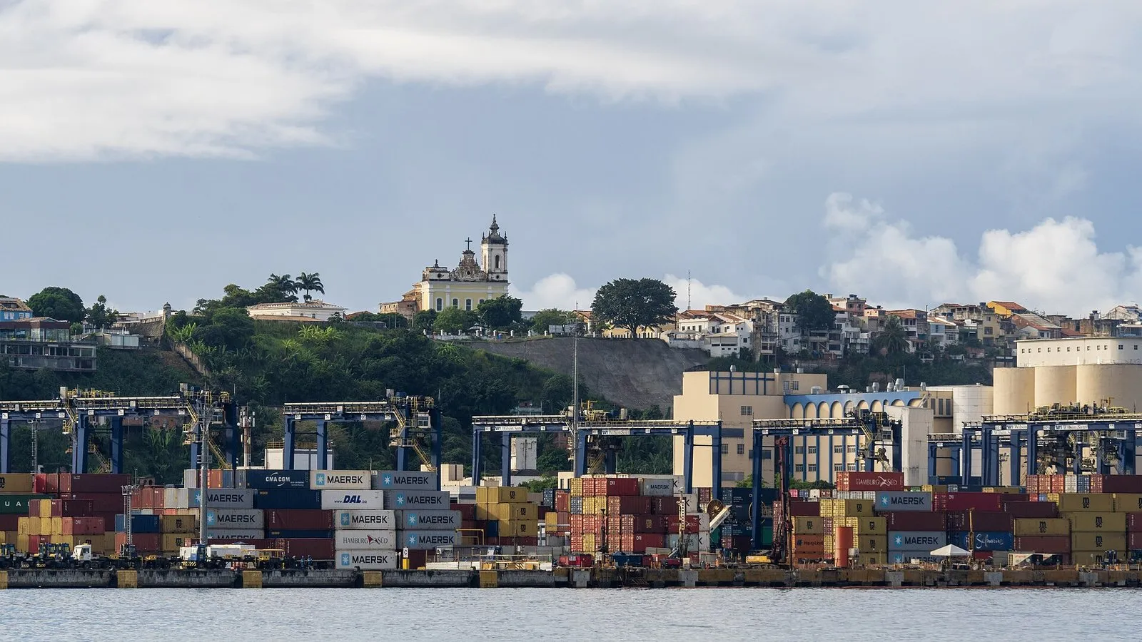 Pilhas de contêineres coloridos e guindastes no terminal de cargas do Porto de Salvador, com casas e uma igreja no alto do morro ao fundo