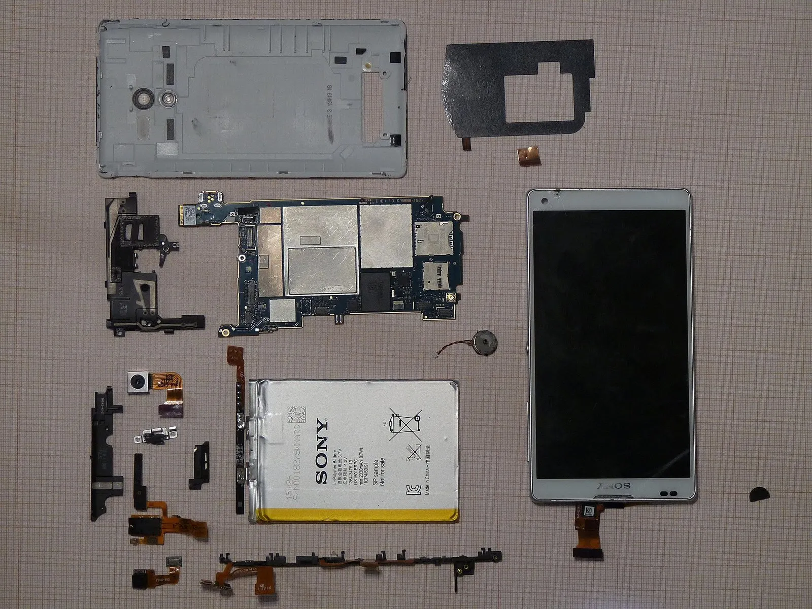 Peças de um smartphone desmontado sobre papel quadriculado: tampa traseira, placa de circuito, bateria, tela, câmeras, motor de vibração e cabos flexíveis