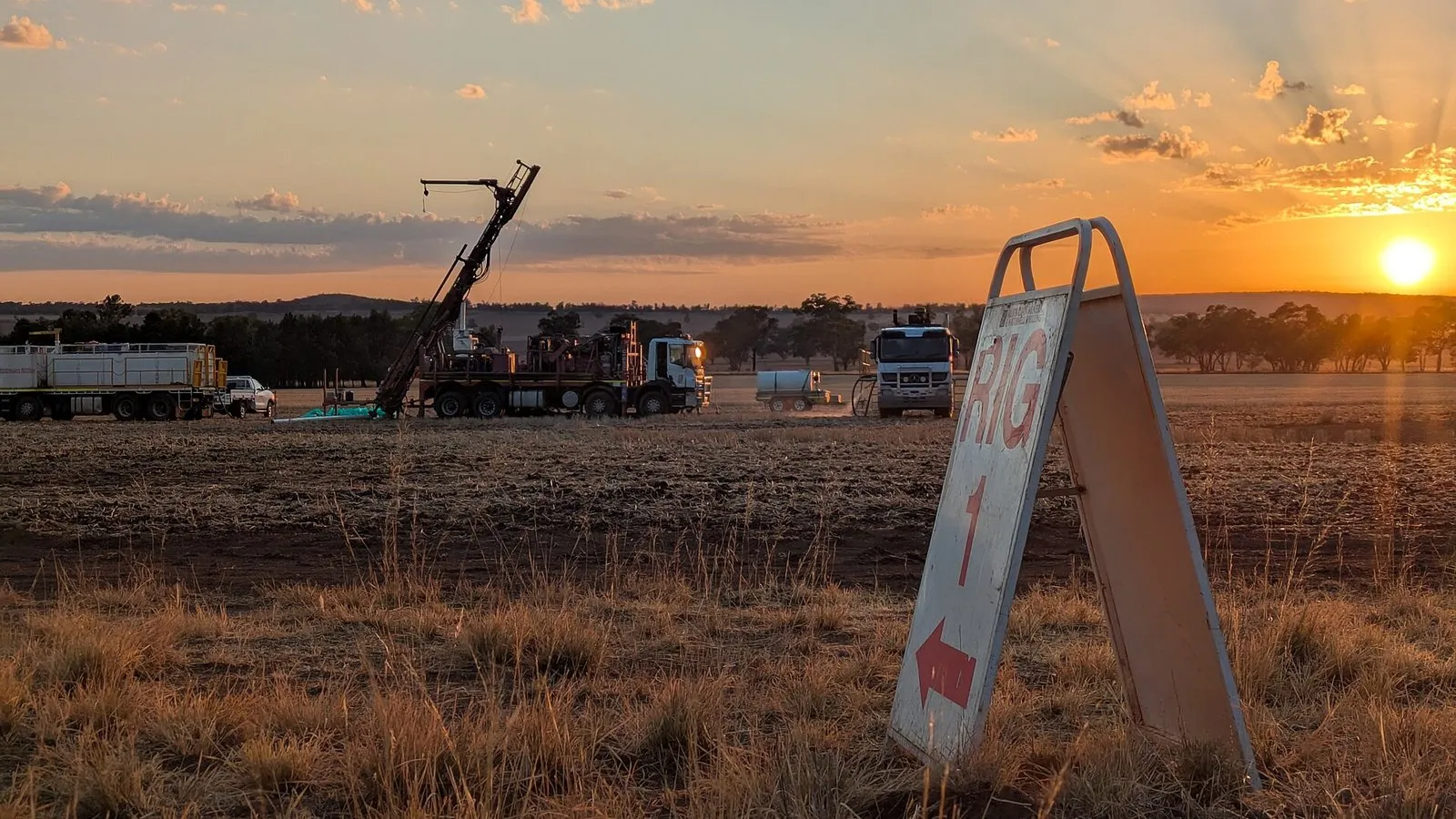 Sonda de perfuração montada em caminhão em um campo aberto ao nascer do sol, com outros caminhões de apoio e uma placa escrita "Rig 1" em primeiro plano