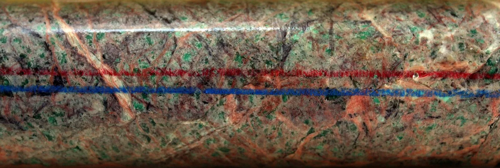 Close de um cilindro de rocha esverdeada com veios avermelhados, com uma linha vermelha e uma azul pintadas ao longo do comprimento